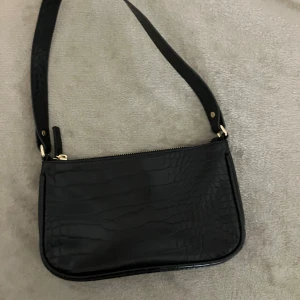 Svart axelväska med krokodilmönster - Snygg svart axelväska med krokodilmönster och dragkedja. Perfekt för att ge en stilren touch till din outfit. Väskan har en bred axelrem för extra komfort. Köpt för 200 säljer för 65.
