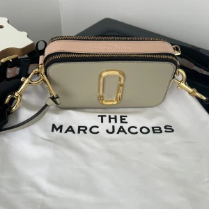 Beige Marc Jacobs Snapshot - Snygg beige axelväska från Marc Jacobs med gulddetaljer och en justerbar axelrem. Väskan har en stilren design med en dragkedja upptill och det ikoniska Marc Jacobs-emblemet på framsidan. Perfekt för att bära dina nödvändigheter med stil.