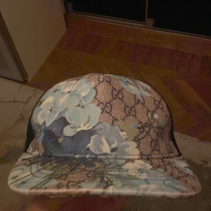 Blommig keps från Gucci - Snygg keps från Gucci med ett blommigt mönster i blått och vitt på en brun bakgrund med logotypmönster. Kepsen har ett nät på baksidan för ventilation och justerbar passform.