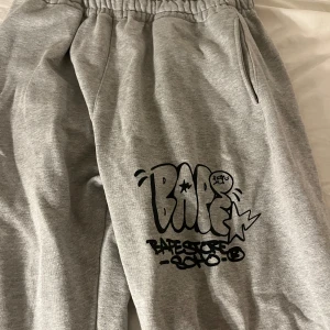 Grå mjukisbyxor från Bape - Säljer ett par grå mjukisbyxor från Bape med elastisk midja och coolt tryck på benet. Byxorna har fickor och en avslappnad passform, perfekta för en skön outfit. Pris kan diskuteras 