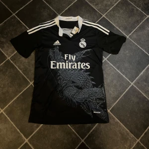 Svart Real Madrid fotbollströja - Snygg svart Real Madrid fotbollströja från Adidas med Fly Emirates som sponsor. Tröjan har ett unikt drakmönster på framsidan och klassiska vita Adidas-ränder på axlarna. Kommer med prislapp på helt ny. Mitt pris 200kr