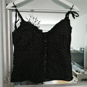 Svart prickig linnetopp - Snygg svart linnetopp med vita prickar och knytband vid axlarna. Toppen har knappar framtill och en smickrande passform. Perfekt för en somrig look!