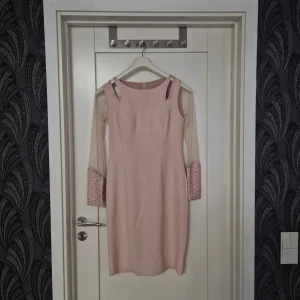Ljusrosa klänning - Elegant ljusrosa klänning med långa transparenta ärmar och glittriga detaljer vid ärmsluten. Klänningen har en figurnära passform och en diskret dragkedja i ryggen. Perfekt för speciella tillfällen.