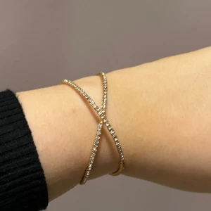 Guldarmband med glittriga detaljer - Elegant guldarmband med ett korsande design och glittriga detaljer. Perfekt för att ge en touch av glamour till din outfit. Justerbar passform för att sitta bekvämt på handleden.