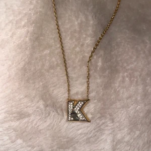 Karl Lagerfeldt halsband - Elegant guldhalsband med ett hängsmycke i form av bokstaven K, dekorerat med glittrande stenar. Perfekt för att lägga till en personlig touch till din stil.