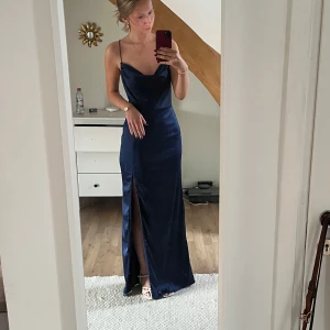 Marinblå balklänning  - Elegant mörkblå långklänning med smala axelband och djup slits framtill. Klänningen har en öppen rygg med korsade band, vilket ger en stilren och sofistikerad look. Perfekt för speciella tillfällen. Endast använd en gång, på min bal 