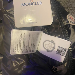 Svart pufferjacka från Moncler - Snygg svart pufferjacka från Moncler med glansig finish. Jackan har en dragkedja framtill och praktiska fickor med dragkedjor. Den har en hög krage och en avtagbar huva för extra skydd mot kyla. Perfekt för kalla dagar.