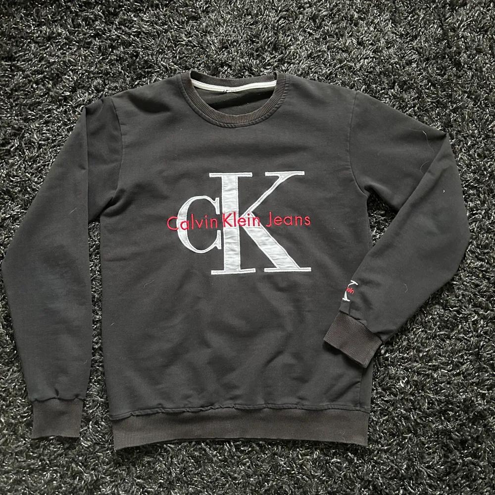 Snygg svart sweatshirt från Calvin Klein med det ikoniska CK-logotypen broderad i vitt och rött på bröstet. Tröjan har långa ärmar och ribbade muddar vid ärmslut och nederkant. Perfekt för en stilren och avslappnad look.. Neuletakit & Villapaidat.