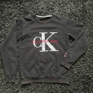 Svart sweatshirt från Calvin Klein - Snygg svart sweatshirt från Calvin Klein med det ikoniska CK-logotypen broderad i vitt och rött på bröstet. Tröjan har långa ärmar och ribbade muddar vid ärmslut och nederkant. Perfekt för en stilren och avslappnad look.