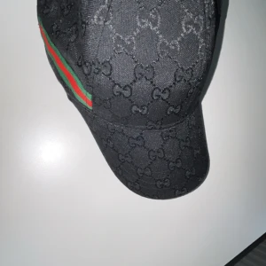 Svart keps med mönster - Snygg svart keps med diskret mönster och justerbar rem baktill. Kepsen har en klassisk design med en liten skärm och en grön och röd randdetalj på sidan.