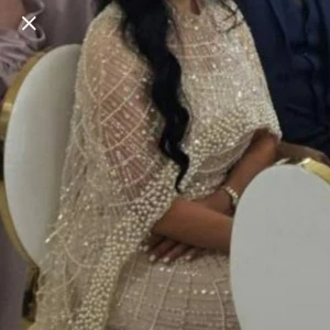 Festkläning med pärlor - Elegant långklänning i champagnefärg med glittrande pärldetaljer. Klänningen har en figurnära passform och är täckt av ett nätmönster av pärlor som ger en lyxig känsla. Perfekt för speciella tillfällen.