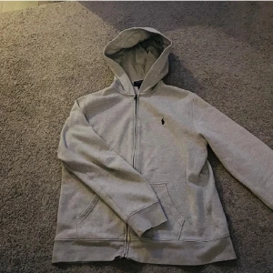 Grå hoodie från Polo Ralph Lauren - Säljer en stilren grå hoodie från Polo Ralph Lauren med dragkedja och klassisk logga på bröstet. Perfekt för en avslappnad look. Den har en bekväm passform och praktiska fickor framtill.