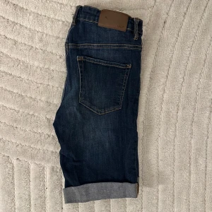 Mörkblå jeansshorts från H&M - Snygga mörkblå jeansshorts från H&M:s L.O.G.G. kollektion. De har en klassisk femficksdesign och uppvikta benslut för en avslappnad look. Perfekta för en casual stil. Använda endast några fåtal gånger