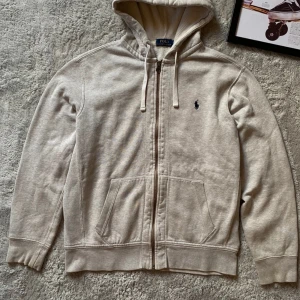 Beige hoodie från Polo Ralph Lauren - Säljer en snygg och bekväm beige hoodie från Polo Ralph Lauren. Den har en dragkedja framtill och en klassisk broderad logga på bröstet. Perfekt för en avslappnad stil och passar till alla tillfällen. Den är i bra skick och redo för en ny ägare!