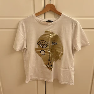 Vit t-shirt med guldtryck från Zara - Säljer en snygg vit t-shirt från Zara med ett coolt guldtryck av ett abstrakt ansikte på framsidan. T-shirten är kortärmad och i bomull, perfekt för en avslappnad stil. Passar både till vardags och festliga tillfällen! Praktiskt sett tagen ny, använd 1 gång.