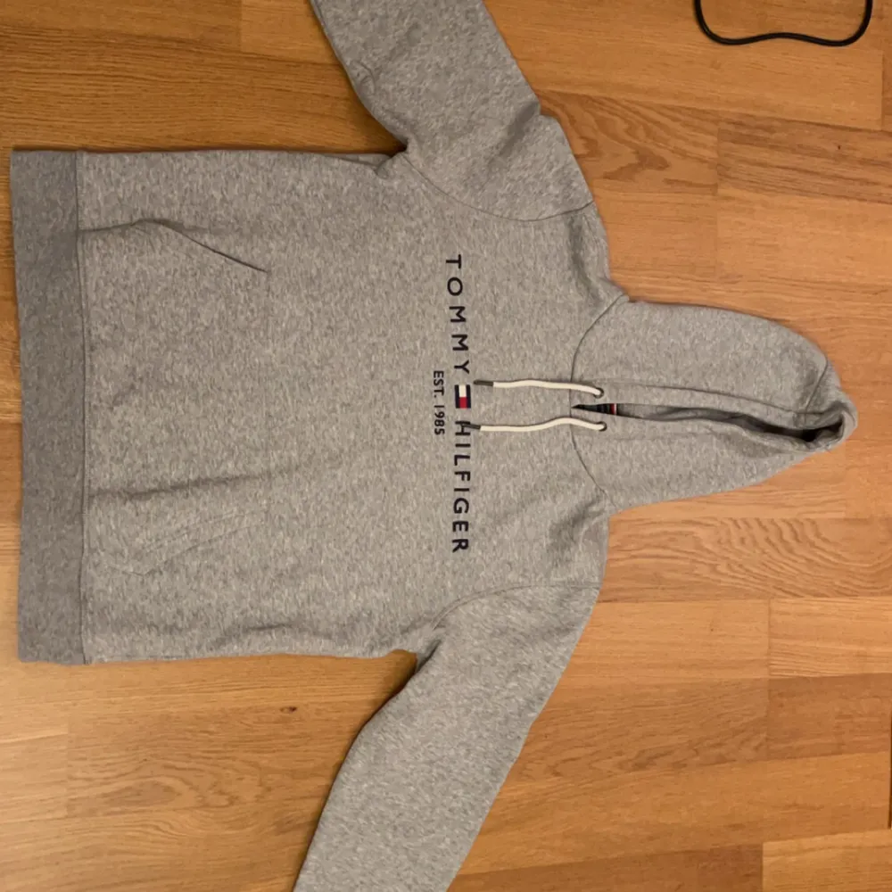 Säljer en snygg och bekväm grå hoodie från Tommy Hilfiger. Den är i storlek M och gjord av ekologisk bomull. Hoodien har en klassisk design med logotypen 'Tommy Hilfiger EST. 1985' broderad på framsidan. Perfekt för en avslappnad stil och passar till alla tillfällen. Den har en känguruficka och justerbar huva med snören.. Tröjor & Koftor.