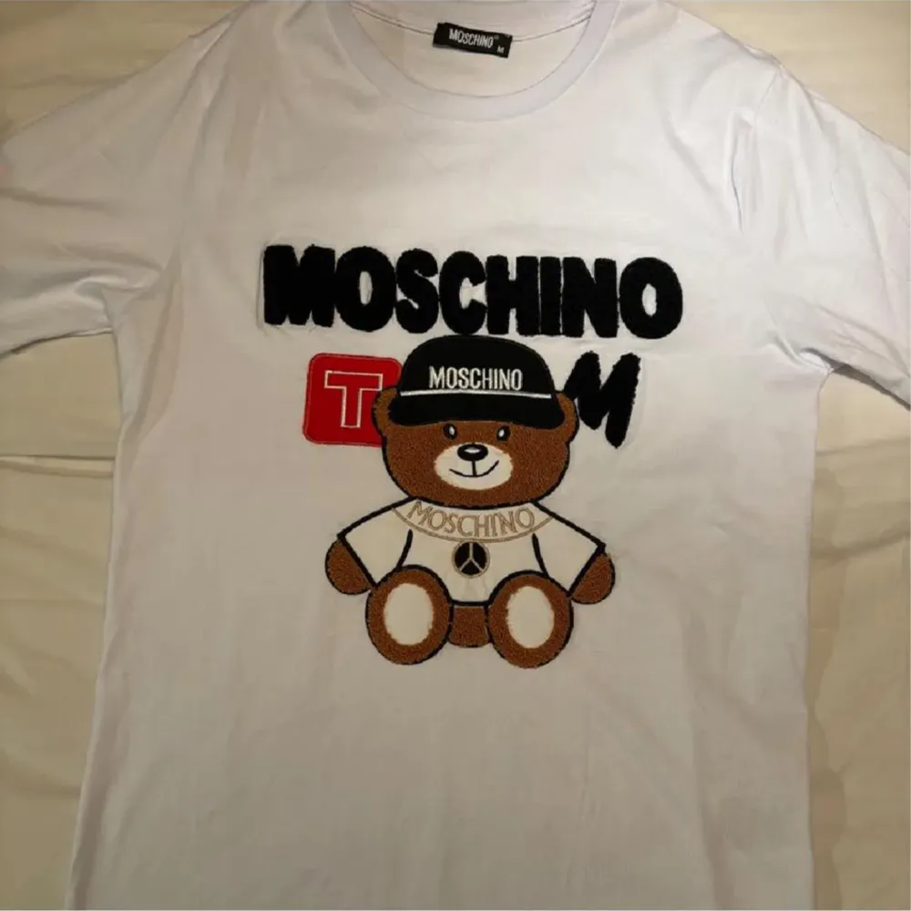 Säljer en supercool vit t-shirt från Moschino med ett stort tryck av en nallebjörn som bär en Moschino-keps. T-shirten har korta ärmar och är i bomull, perfekt för en avslappnad och trendig look. Passar perfekt till både jeans och shorts!. T-shirts.