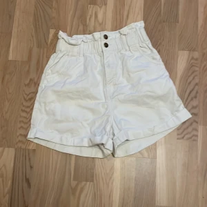 Vita bomullsshorts med hög midja - Säljer ett par vita shorts i storlek 36, tillverkade av 100% bomull. De är ganska mellan midja med elastisk resår och två knappar framtill. Perfekta för sommardagar och kan enkelt matchas med en t-shirt eller blus. Använda men i bra skick!