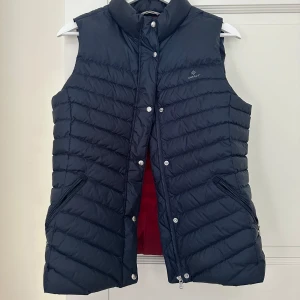 Marinblå dunväst från GANT - Säljer en snygg och stilren marinblå dunväst från GANT. Västen har en quiltad design med dragkedja och knappar framtill. Insidan är fodrad med en cool röd färg som ger en fin kontrast. Barnstorlek M / 160. nypris är cirka 1600kr