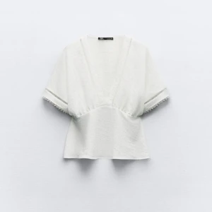 Vit blus med spetsdetaljer - Säljer en superfin vit blus med v-ringning och spetsdetaljer vid ärmslut och hals. Den har korta ärmar och en lös passform som gör den perfekt för sommardagar. Blusen är gjord i ett lätt och luftigt material, vilket gör den bekväm att bära hela dagen. Den är slutsåld på Zara. Nypris 379.