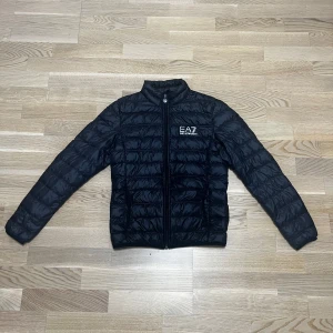 Svart pufferjacka från Emporio Armani - Säljer en snygg svart jacka från Emporio Armani EA7. Jackan är i mycket bra skick och har en dragkedja framtill. Perfekt för lite extra värme, med långärmade ärmar och quiltad design för mera värme. Den är både stilren och funktionell! Köpte den av min kusin för att det inte passade han och nu säljer jag den för att tömma garderoben. Storleken är (S)