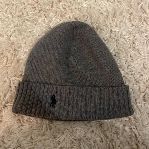 Grå stickad mössa från Polo Ralph Lauren - Säljer en snygg grå stickad mössa från Polo Ralph Lauren. Den är i merinoull och har en broderad logga framtill. Perfekt för kalla dagar och ger en stilren look. Mössan är i mycket bra skick och passar de flesta huvudstorlekar.