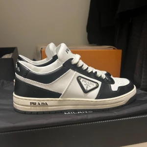 Prada Sneakers - Säljer ett par snygga svartvita sneakers från Prada i mycket bra skick. De har en klassisk design med Pradas logga på sidan och är perfekta för både vardag och fest. Skorna har snörning och en bekväm sula som ger bra stöd. Perfekta för dig som vill ha stil och komfort i ett! Kvitto finns för seriösa köpare. 