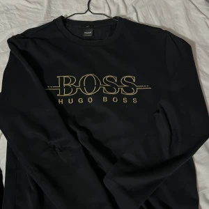 Svart tröja från Hugo Boss - Säljer en stilren svart tröja från Hugo Boss med deras ikoniska logga i guld på bröstet. Tröjan har långa ärmar och en klassisk rund halsringning. Perfekt för både vardag och fest. Passar till allt och ger en lyxig touch till din outfit.tröjan har dessutom slitna märken på armbågarna men man märker inte så mycket pågrund av att det är mörkt där.