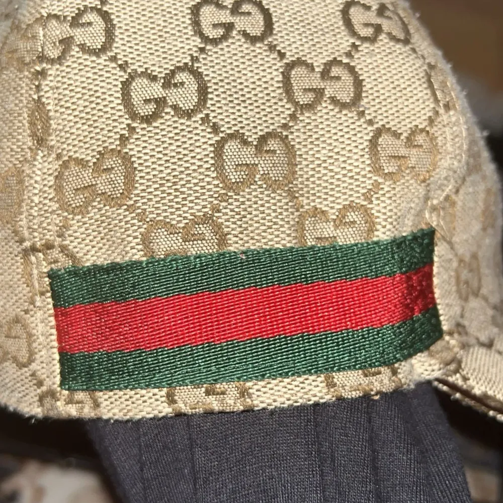 Säljer en snygg beige keps från Gucci med deras ikoniska GG-mönster. Kepsen har en klassisk design med en röd och grön randdetalj på sidan. Perfekt för att ge din outfit en lyxig touch. Tillverkad i Italien och i mycket bra skick.. Accessoarer.