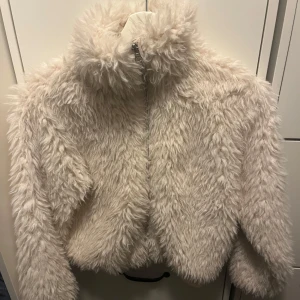Beige pälsjacka med dragkedja - Supermysig beige teddyjacka som är perfekt för kyliga dagar. Den har en dragkedja framtill och är helfodrad för extra värme. Jackan är långärmad och har en fluffig look som ger en trendig stil. Perfekt för höst och vinter!
