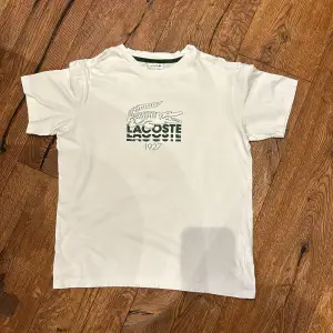 Säljer en klassisk vit t-shirt från Lacoste i storlek 14. Den har det ikoniska krokodilmärket broderat på framsidan och en grön rand vid kragen. Perfekt för en avslappnad stil eller sommarens äventyr. T-shirten är i bra skick och redo för en ny ägare!