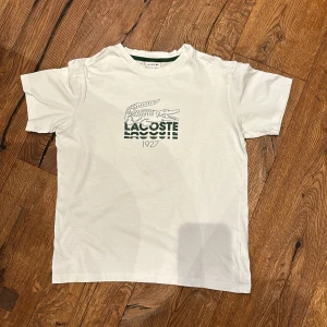 Vit t-shirt från Lacoste - Säljer en klassisk vit t-shirt från Lacoste i storlek 14. Den har det ikoniska krokodilmärket broderat på framsidan och en grön rand vid kragen. Perfekt för en avslappnad stil eller sommarens äventyr. T-shirten är i bra skick och redo för en ny ägare!