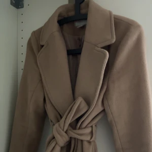 Beige kappa med knytband - Säljer en superfin beige kappa i ull med knytband i midjan. Perfekt för höst och vår! Den har långa ärmar och en klassisk krage. Kappan är i mycket bra skick och ger en elegant look. Passar både till vardags och fest.