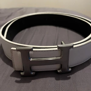 Vitt skärp med silverfärgat spänne - Säljer ett stilrent vitt skärp i skinn med ett stort silverfärgat H-format spänne. Perfekt för att ge en touch till vilken outfit som helst. Skärpet är i mycket bra skick. Storlek 110.