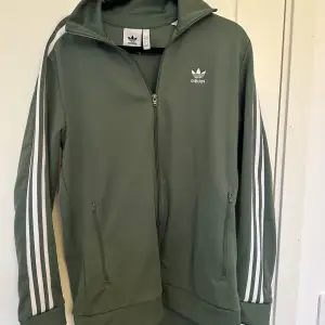 Säljer en snygg grön träningsjacka från Adidas i storlek L. Jackan har de klassiska tre vita ränderna längs ärmarna och en dragkedja framtill. Perfekt för träning eller som en casual vardagslook. Den är i bra skick och redo för nya äventyr! 💚