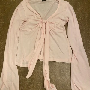 Ljusrosa långärmad topp med knyt - Säljer en söt ljusrosa topp med långa ärmar och knyt framtill. Den är i ett mjukt och bekvämt material, vilket gör den idealisk för både vardag och festliga tillfällen. Toppen har en v-ringad design som ger en feminin touch. 