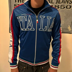 Blå track jacket med Italiens flagga - Säljer en snygg blå track jacket med texten 'ITALIA' på framsidan och Italiens flagga på kragen. Jackan har dragkedja och långa ärmar med ränder i Italiens färger längs sidorna. Perfekt för sportiga tillfällen eller som en stilren vardagsjacka.