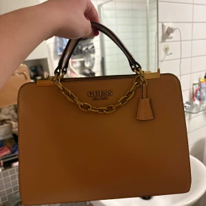 Brun handväska från Guess - Snygg brun handväska från Guess i mycket bra skick. Väskan har en elegant kedjedetalj och dragkedja för säker förvaring. Perfekt för både vardag och festliga tillfällen. Stilren design som passar till det mesta!