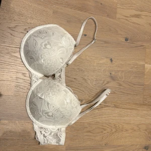 Vit spets-bh - Säljer en elegant vit bh med spetsdetaljer. Den har justerbara axelband och en klassisk hakknäppning bak. Perfekt för en romantisk look eller till vardags under en blus. Spetsen ger en feminin touch och den är både bekväm och stilren. Storlek 75 D🩷