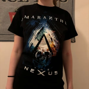 Svart band-t-shirt med tryck - Säljer en svart band-t-shirt från Amaranthe med coolt tryck på framsidan och turnédatum på baksidan. Perfekt för konserter eller vardagsstil. T-shirten är kortärmad och i bomull, vilket gör den bekväm att bära. Passar bra till jeans eller shorts för en avslappnad look. The nexus