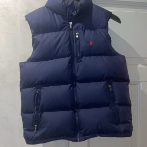 Blå Ralph lauren väst - Säljer en snygg mörkblå dunväst från Ralph Lauren. Västen har en dragkedja framtill och två praktiska fickor med dragkedjor. Perfekt för kyliga dagar och ger en stilren look. Den är i mycket bra skick och passar perfekt för höst och vinter. ❄️ Hör gärna av dig om du har frågor! Storlek 18-20 år eller barnstorlek:XL vuxen storlek: M