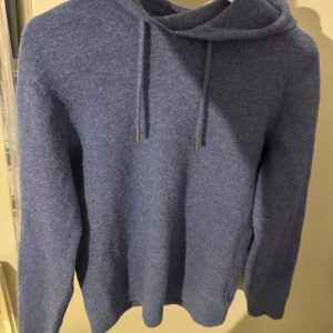 Blå hoodie i kashmir - Säljer en supermjuk och stilren blå hoodie i merinoull i storlek L men sitter som än S . Perfekt för kyliga dagar med sin varma och sköna känsla. Den har en klassisk design med dragsko i huvan och långa ärmar.