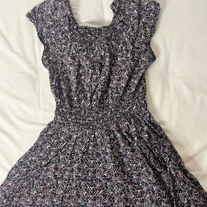 Blommig klänning med volangdetaljer - Söt blommig klänning i mörkblått med små vita och rosa blommor. Den har volangdetaljer vid ärmarna och en smockad midja för en bekväm passform. Perfekt för vår och sommar!