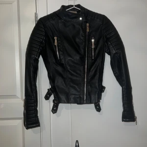 Svart skinnjacka med dragkedjor - Säljer en snygg svart skinnjacka i mycket bra skick. Den har coola dragkedjor och spännen på sidorna för en edgy look. Perfekt för höst och vår, och funkar lika bra till fest som till vardags. Jackan har långa ärmar och en tight passform. Ett måste för garderoben!