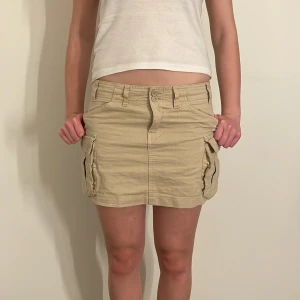 Beige cargokjol med fickor - Säljer en superfin beige cargokjol. Kjolen har en klassisk design med bälteshällor och knappstängning framtill. Från Brandy Melville 