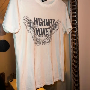 Vit t-shirt med tryck - Säljer en vit t-shirt med coolt tryck 'Highway Honey Tour of 1979' och vingar. Perfekt för en avslappnad stil eller konsert. T-shirten är kortärmad och i bra skick. Passar perfekt till jeans eller shorts!