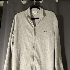 Grå kofta från Lacoste - Säljer en stilren grå kofta från Lacoste i regular fit. Den har en dragkedja framtill och det klassiska krokodilmärket på bröstet. Perfekt för både vardag och lite finare tillfällen. Superbekväm och lätt att matcha med andra plagg! Ny 1200kr