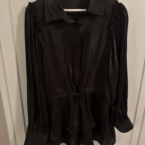 Svart glansig blus - Svart glansig blus med volanger nertill. I storlek L. Som ny i skicket. I köpt för 1299:-