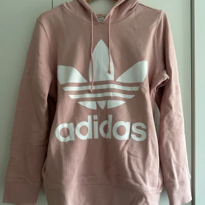 Rosa hoodie från Adidas - Säljer en superfin rosa hoodie från Adidas i storlek 36. Den har en stor vit logga på framsidan samt fickor. Perfekt för höst och vår! Den är i mycket bra skick och har en skön passform med långa ärmar och luva