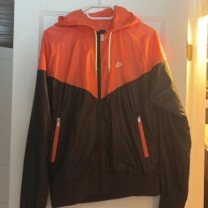 Svart och orange vindjacka från Nike - Säljer en snygg svart och orange vindjacka från Nike. Jackan har dragkedja fram och justerbar huva med snören. Använd därav priset. Storleken är oklar men skulle säga att den passar S-M